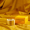 Supercharge scented100% natural wax candle 110g in pure brass container next to bright yellow candle box with closed lid. Front.