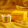 Supercharge scented100% natural wax candle 110g in pure brass container next to bright yellow candle box with closed lid. Front.